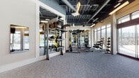 This fitness center is designed to accommodate a wide range of workout styles across several distinct zones. The main gym area features soaring exposed ceilings with visible ductwork and steel framing painted in dark tones, contributing to an industrial-modern aesthetic. Bold yellow and white chevron graphics accent the walls alongside stone-textured surfaces and large "GYM" lettering, creating an energetic and motivating atmosphere. The flooring throughout the primary cardio and strength areas consists of durable, textured carpet tile, while a dedicated training zone features rich wood-look flooring for functional and group fitness activities.

The cardio section is lined with treadmills and stair climbers positioned in front of expansive floor-to-ceiling windows and glass doors that look directly onto an outdoor pool area, providing abundant natural light and scenic views during workouts. The strength training area is well-appointed with a full rack of dumbbells, cable machines, TRX suspension trainers, medicine balls, and multi-functional cable systems, all reflected in large wall-mounted mirrors. A designated Training Zone room offers spin bikes, Pilates equipment, and additional functional training tools. The space also includes staircase access to an upper mezzanine level, adding to the overall vertical scale and open feel of the facility.