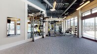 This fitness center is designed to accommodate a wide range of workout styles across several distinct zones. The main gym area features soaring exposed ceilings with visible ductwork and steel framing painted in dark tones, contributing to an industrial-modern aesthetic. Bold yellow and white chevron graphics accent the walls alongside stone-textured surfaces and large "GYM" lettering, creating an energetic and motivating atmosphere. The flooring throughout the primary cardio and strength areas consists of durable, textured carpet tile, while a dedicated training zone features rich wood-look flooring for functional and group fitness activities.

The cardio section is lined with treadmills and stair climbers positioned in front of expansive floor-to-ceiling windows and glass doors that look directly onto an outdoor pool area, providing abundant natural light and scenic views during workouts. The strength training area is well-appointed with a full rack of dumbbells, cable machines, TRX suspension trainers, medicine balls, and multi-functional cable systems, all reflected in large wall-mounted mirrors. A designated Training Zone room offers spin bikes, Pilates equipment, and additional functional training tools. The space also includes staircase access to an upper mezzanine level, adding to the overall vertical scale and open feel of the facility.