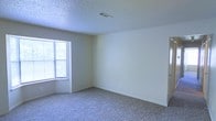 This single-floor space presents a straightforward and functional layout with a clean, neutral aesthetic throughout. The interior is finished with soft, wall-to-wall carpeting that adds warmth underfoot and contributes to a quiet, comfortable atmosphere. Walls are painted in a light, neutral tone that reflects natural light and provides a versatile backdrop for any décor style. A notable architectural feature is the recessed bay-style window, which draws in generous amounts of natural light and creates a bright, open feel within the room. The textured popcorn ceiling adds a classic character to the space, while a ceiling-mounted HVAC vent indicates a centralized climate control system for year-round comfort. Electrical outlets are conveniently positioned along the baseboards, ensuring practical usability throughout. The overall ambiance is calm and understated, with an open feel that lends itself well to a variety of furniture arrangements and uses. The space is presented in a vacant, move-in-ready condition, offering a blank canvas for residents or tenants to customize to their needs and preferences.