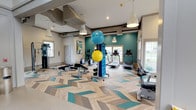 Located at 510 Stevens Ave SW in Renton, Washington, this fitness center offers a thoughtfully designed, single-floor workout facility built to accommodate a wide range of exercise needs. The space immediately draws the eye with its striking chevron-patterned flooring, which blends natural wood tones with teal accents, creating a dynamic and energizing atmosphere throughout.

The interior features a bold teal accent wall that adds vibrancy to the space, complemented by crisp white walls and warm pendant lighting that hangs from the raised ceiling. Recessed lighting and large windows along the perimeter flood the room with natural light, providing views of the surrounding outdoor greenery and contributing to an open, airy feel.

The fitness center is well-equipped with a variety of cardio machines, including treadmills and stationary bikes, positioned near the windows to take advantage of natural light during workouts. A dedicated free weight and functional training area includes adjustable benches, dumbbells, and stability balls organized on a centrally located rack. The layout is spacious and thoughtfully arranged to allow ease of movement between equipment zones. The overall ambiance is clean, modern, and inviting, making it a well-rounded facility suitable for residents and members seeking a comprehensive fitness experience.