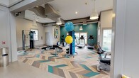 Located at 510 Stevens Ave SW in Renton, Washington, this fitness center offers a thoughtfully designed, single-floor workout facility built to accommodate a wide range of exercise needs. The space immediately draws the eye with its striking chevron-patterned flooring, which blends natural wood tones with teal accents, creating a dynamic and energizing atmosphere throughout.

The interior features a bold teal accent wall that adds vibrancy to the space, complemented by crisp white walls and warm pendant lighting that hangs from the raised ceiling. Recessed lighting and large windows along the perimeter flood the room with natural light, providing views of the surrounding outdoor greenery and contributing to an open, airy feel.

The fitness center is well-equipped with a variety of cardio machines, including treadmills and stationary bikes, positioned near the windows to take advantage of natural light during workouts. A dedicated free weight and functional training area includes adjustable benches, dumbbells, and stability balls organized on a centrally located rack. The layout is spacious and thoughtfully arranged to allow ease of movement between equipment zones. The overall ambiance is clean, modern, and inviting, making it a well-rounded facility suitable for residents and members seeking a comprehensive fitness experience.