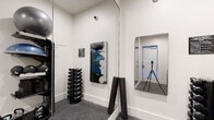 This single-level fitness room is thoughtfully designed to maximize functionality within a compact footprint. The space features clean white walls and durable speckled rubber flooring, creating a practical and hygienic environment suited for a variety of workout routines.

A standout feature of the room is the pair of Echelon Reflect smart fitness mirrors mounted on the wall, which enable interactive, instructor-led workouts directly from the space. A full-length wall mirror further enhances the sense of openness while allowing users to monitor their form during exercise. A camera on a tripod is positioned in the room, complementing the smart mirror setup for virtual training sessions.

Equipment is neatly organized throughout the space. A tiered dumbbell rack holds a graduated set of weights, while wall-mounted shelving accommodates balance trainers, medicine balls, and other functional fitness accessories. Foam rollers and kettlebells are also present, supporting a well-rounded training experience.

The entrance area features a keypad-secured door with a stainless steel waste receptacle nearby, maintaining a clean and organized entry point. A security camera is mounted near the ceiling, ensuring safety throughout the space. The overall ambiance is modern, clean, and purposefully utilitarian.