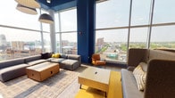 The Sky Lounge is a thoughtfully designed, elevated communal space that seamlessly blends relaxation, collaboration, and striking urban views. The lounge is anchored by an expansive wall of floor-to-ceiling windows that flood the interior with natural light and frame sweeping panoramic views of the surrounding cityscape and tree-lined neighborhoods beyond.

The seating area is outfitted with a curated mix of contemporary furniture, including modular gray sofas accented with mustard yellow and striped throw pillows, mid-century-inspired cognac leather armchairs, and high-back lounge chairs — all arranged atop layered area rugs in yellow and muted geometric patterns. Bold navy blue accent walls provide a striking contrast to the light-filled atmosphere, reinforcing the space's modern, energetic aesthetic.

Transitioning into the collaborative zone, a large communal high-top table on casters serves as a flexible centerpiece, surrounded by sleek white bar stools and illuminated by oversized pendant light fixtures with an industrial character. Exposed ductwork at the ceiling adds an urban loft-like quality. Warm wood wall paneling and curated wall art contribute texture and personality to the space.

Additional rooms visible from the main area include enclosed glass-walled rooms ideal for private meetings or focused work. Together, these elements make the Sky Lounge a dynamic, multi-purpose destination suited for socializing, working, or simply taking in the view.
