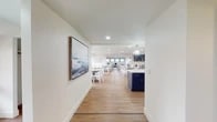 This single-floor communal space is thoughtfully designed with a coastal aesthetic that blends functionality and style throughout. Warm hardwood flooring runs through the main corridor, kitchen, and dining areas, creating a cohesive flow from one zone to the next. A striking large-scale artwork greets visitors upon entry, setting an inviting tone.

The kitchen is a standout feature, showcasing bold navy lower cabinetry paired with crisp white upper cabinets, gold hardware, quartz countertops, a decorative tile backsplash, and stainless steel appliances including a range and over-the-range microwave. A navy island adds additional workspace and visual impact.

Multiple dining areas are arranged throughout the open layout, furnished with round tables and white cross-back chairs. Shiplap accent walls lend a clean, nautical character to several zones, including a dedicated lounge area featuring a wall-mounted television, herringbone wood detail, and plush striped armchairs. An adjacent seating area with large panoramic windows offers sweeping outdoor views and abundant natural light. A well-appointed accessible restroom with herringbone tile completes the space.