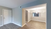 This single-story space offers a clean, open layout connecting the living and dining areas in a straightforward, functional design. The flooring throughout is wood-look planking in a warm tone, providing a cohesive and durable surface that flows seamlessly between rooms. Walls are painted in a soft blue-gray hue in the main living area, transitioning to neutral beige tones in the adjacent dining space, creating a subtle visual distinction between the two zones. A textured ceiling adds modest dimension overhead, while white baseboards provide crisp contrast along the lower walls. The dining area is illuminated by a pendant chandelier with a classic tiered design, lending a touch of elegance to the space. A large mirror in the dining area reflects natural light from a nearby window, enhancing the sense of openness and brightness. Overhead ventilation is provided by a ceiling-mounted HVAC register. A doorway visible from the main area leads to an additional room or hallway, suggesting further functional spaces beyond the main living zone. The overall ambiance is neutral, tidy, and move-in ready, suitable for a variety of interior styles.