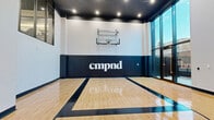 Located at 632 Newark Avenue in Jersey City, NJ, this indoor basketball court is part of the CMPND facility and offers a well-appointed athletic space designed for serious play. The court features a glossy, regulation-marked hardwood floor with bold black lane lines and a three-point arc, providing a professional playing surface. A wall-mounted glass backboard and hoop anchor the main end of the court, backed by navy blue padded wall panels bearing the CMPND logo for both safety and branding. The space is defined by its dramatic double-height ceilings, which create an open, airy atmosphere essential for full court play. Recessed lighting throughout the dark-painted ceiling ensures bright, even illumination across the entire floor. A striking floor-to-ceiling glass wall and large glass entry doors on one side of the court allow abundant natural light to flood the space while offering views of the surrounding outdoor area. An interior staircase and a mezzanine-level overlook with glass-paneled windows add a dynamic vertical element, allowing views down onto the court from above. The clean white walls and dark accents give the space a modern, polished aesthetic, making it a standout athletic amenity within the broader CMPND complex.