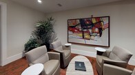 This thoughtfully designed single-floor office suite combines functionality with sophisticated aesthetics. Rich, patterned carpet in warm terracotta tones flows throughout the space, anchoring each area with a cohesive visual identity. The conference room offers a substantial meeting table surrounded by upholstered chairs beneath a striking geometric pendant light fixture, complemented by large windows dressed in bold floral-patterned drapes that allow natural light to filter in. Curated artwork adorns the walls, adding an artistic dimension to the professional setting. A private office area features a striking floor-to-ceiling wood-paneled accent wall in dark tones with horizontal banding, creating a dramatic backdrop for dual workstations equipped with desktop computers. Glass partitions separate this workspace, maintaining visual openness while providing acoustic separation. A dedicated business center offers additional workstations with sleek white desks and modern monitors. The lounge area provides a welcoming retreat with curved upholstered armchairs, small accent tables, lush greenery, and a large-scale abstract artwork as a focal point. Throughout the suite, recessed lighting ensures consistent illumination, while the refined material palette of wood, glass, and neutral tones creates a polished, contemporary professional environment.