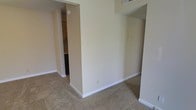 This single-story space features a straightforward, functional layout with a neutral color palette throughout. The main living area is finished with wall-to-wall beige carpeting and cream-colored walls, creating a clean, understated aesthetic. White baseboards provide a crisp contrast along the floor line. A partial wall divides portions of the space, adding subtle architectural definition without fully closing off the floor plan. An open passageway on the left side of the image leads into what appears to be a kitchen area, where cabinetry and countertops are visible. The kitchen appears to have hard flooring, distinguishing it from the carpeted living spaces. Overhead, a ceiling-mounted vent is visible, indicating a central HVAC system. Electrical outlets and switch plates are positioned along the walls at standard heights. The overall ambiance is simple and move-in ready, offering a blank canvas that accommodates a variety of furnishing styles. The connected layout between the living area and kitchen promotes ease of movement throughout the space. Natural light levels are modest based on the image, with interior lighting supplementing the ambient brightness of the rooms.