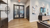 Located at 2945 Brick Church Pike in Nashville, TN, this well-appointed single-floor space offers a cohesive blend of modern design and functional layout across its living, dining, and kitchen areas. The entry welcomes visitors through bold black-framed glass French doors, setting an immediate tone of contemporary sophistication. Warm hardwood-style flooring flows consistently throughout, tying the spaces together seamlessly. The living area serves as a central gathering hub, anchored by a geometric lattice accent wall that frames a mounted flat-screen TV above a sleek wood-and-metal credenza. Plush seating arrangements and curated décor, including sculptural accents and abstract artwork, create an inviting and refined atmosphere. Large windows along the perimeter flood the space with natural light while offering views of the surrounding greenery. The dining area features a substantial natural wood table paired with leather-upholstered chairs and illuminated by a modern candelabra chandelier, complemented by vibrant wall art. A secondary dining nook includes a round wood high-top table with mid-century modern bar chairs and open metal shelving. The kitchen is outfitted with rich dark cabinetry, white subway tile backsplash, ample counter space, and full-size appliances, all brightened by dual windows overlooking the exterior.