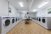 Located at 940 Commissioners Rd in London, Ontario, this shared laundry facility offers a well-organized and functional space designed to accommodate the needs of multiple users efficiently. The room is arranged with rows of modern front-load washing machines and dryers lining both side walls, providing ample capacity for residents. The machines are clearly labeled and accompanied by posted instructional signage to guide users through proper operation.

At the center of the room sits a large, tiled folding table accompanied by a chair, offering a convenient surface for sorting and folding laundry. The flooring throughout consists of durable yellow ceramic tiles, well-suited for a high-traffic utility environment. Overhead fluorescent lighting ensures the space is brightly and evenly lit.

Near the entrance, a Coca-Cola vending machine provides added convenience for users during their visits. A bulletin board on the wall displays notices and information relevant to residents. The room also features a baseboard heater, ensuring comfort during colder months. The neutral grey-and-white walls and clean, practical layout give the space a tidy and functional atmosphere. A security camera is mounted near the ceiling, contributing to the safety and oversight of the facility. Overall, the laundry room is a well-equipped, no-frills communal space designed for practicality and ease of use.