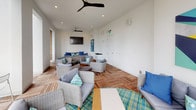 Located at 109 W Fortune St in the heart of downtown Tampa, Florida, this sophisticated lounge space offers a seamless blend of indoor comfort and open-air ambiance. The room features a distinctive curved wall of floor-to-ceiling windows that flood the interior with natural light while framing expansive views of the surrounding cityscape.

Underfoot, warm teak wood deck tiles are laid in a herringbone-inspired pattern, lending a resort-like aesthetic throughout the space. Ceiling fans overhead ensure comfortable airflow, complementing the bright white walls and recessed lighting that keep the atmosphere airy and inviting.

The lounge is furnished with an arrangement of wicker and upholstered chairs and sofas, accented with teal, blue, and aqua throw pillows that echo a coastal color palette. Seating clusters are thoughtfully arranged to encourage both intimate conversations and larger group gatherings. A wall-mounted flat-screen television anchors one end of the room, while vibrant abstract artwork — including a bold blue canvas and a colorful geometric piece — adds visual interest to the clean white walls.

A plaid area rug defines one seating zone, adding texture and warmth to the space. Recycling stations are discreetly integrated, reflecting a commitment to sustainability. Overall, the space exudes a polished, modern coastal atmosphere perfectly suited to its vibrant downtown Tampa location.