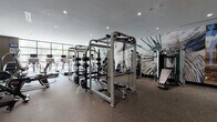 This modern fitness center is designed to support a full range of exercise disciplines within a single, thoughtfully organized space. Large floor-to-ceiling windows along one wall flood the cardio zone with natural light and offer open views of the surrounding streetscape, creating an energizing environment for treadmill and cycling workouts. A row of commercial-grade treadmills with individual display screens lines this windowed wall, complemented by stationary bikes and a rowing machine nearby. Full-length mirrors expand the sense of space and allow users to monitor their form throughout strength training sessions. The strength zone is anchored by multiple cable and power rack systems, accommodating a wide variety of resistance exercises. A dedicated free weights area features a comprehensive dumbbell rack alongside flat benches and a wall-mounted flat-screen television. Functional training needs are met with stability balls, medicine balls, TRX suspension systems, and additional accessories displayed on a rack set against a vibrant, colorful mural. Throughout the facility, bold and abstract murals depicting athletic silhouettes add an artistic energy to the space. Recessed lighting ensures consistent illumination, while durable, dark-toned flooring ties the design together. A water fountain station and convenient elevator access are located just outside the gym entrance.