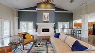 Located at 135 Westfield Ct in Clarksville, Tennessee, this single-story community clubhouse offers a well-rounded amenity facility designed for comfort and functionality. The centerpiece of the space is a grand lounge area showcasing soaring vaulted ceilings, a striking travertine fireplace with an ornate white mantel, and rich hardwood flooring. Deep slate-blue walls contrast elegantly with white wainscoting and crown molding throughout, creating a refined yet welcoming atmosphere. Large windows flood the lounge with natural light and provide views of the lush surrounding greenery. A chandelier overhead adds a touch of classic sophistication to the seating area. Adjacent to the lounge is a generously sized full kitchen outfitted with warm cherry wood cabinetry, ample counter space, a double sink, and modern appliances — ideal for community gatherings and events. The fitness wing is expansive and well-equipped, featuring cardio machines including treadmills and stair climbers, a full rack system, free weights, and mirrored walls. A dedicated yoga and stretching studio with light aqua walls, floor-to-ceiling mirrors, and exercise mat storage complements the main gym area. Additional amenities include a package room with mailbox lockers and a laundry room with multiple full-size washers and dryers.
