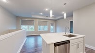 This contemporary two-floor residence is thoughtfully designed with clean lines and modern finishes throughout. The main living level showcases an open-concept layout where the kitchen flows seamlessly into the living area, creating an inviting and functional space. The kitchen is a standout feature, equipped with stainless steel appliances including a refrigerator, range, microwave, and dishwasher. White shaker-style cabinetry pairs elegantly with white quartz countertops and a classic subway tile backsplash. A central island with a built-in sink serves as both a prep area and a gathering point, illuminated by stylish pendant lighting above. Durable hardwood-style flooring runs throughout the main living areas, complemented by neutral warm-toned walls. The living area benefits from tray ceilings that add architectural depth, and multiple windows dressed with roller shades invite natural light while offering views of the surrounding neighborhood. A private balcony door extends the living space outdoors. The bedrooms are generously proportioned and well-lit, each featuring the same quality flooring. The bathroom is cleanly finished with a full soaking tub and shower combination, white tile surround, and modern chrome fixtures. The lower level includes a spacious attached garage with an automatic opener, offering ample storage and parking.