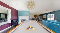Located at 5095 E Donald Ave in Denver, CO, this community room offers a well-appointed, single-floor gathering space designed to accommodate a variety of social and recreational activities. The space is divided into two distinct yet seamlessly connected areas, each thoughtfully designed with modern finishes and vibrant accent colors throughout.

The main lounge area is anchored by a decorative fireplace framed by an elegant white mantel and mosaic tile surround, set against a rich, patterned plum accent wall. A billiards table serves as a central focal point, complemented by plush teal velvet seating and a statement floral chandelier overhead. Large floor-to-ceiling sliding glass doors flood the space with natural light and provide direct views of the surrounding outdoor greenery. A wall-mounted flat-screen TV and bar-height seating add to the room's entertainment versatility.

The fully equipped kitchen features contemporary dark wood cabinetry, stainless steel appliances, white quartz countertops, and a breakfast bar with cushioned stools — ideal for hosting events and gatherings. A coffee station adds a welcoming touch.

The adjacent reception area greets visitors with striking arched wall panels clad in textured blue wallcovering, a geometric pendant light, hardwood-style flooring, and a staffed front desk, establishing a polished and professional atmosphere throughout.