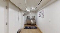 Located at 5764 Stevens Forest Rd in Columbia, MD, this single-floor hallway space is purpose-built as a pet grooming and wash station. The layout is narrow and efficient, with light wood-look flooring running the length of the corridor, creating a clean and easy-to-maintain surface. The centerpiece of the space is a professional-grade stainless steel dog bathing tub, complete with a built-in faucet, handheld sprayer, and a grated floor panel for drainage — ideal for pet washing needs. A playful paw-print mat is placed in front of the tub, adding a welcoming touch. The walls are painted white, contributing to a bright and sanitary atmosphere, and are accented with black-and-white pet-themed artwork. Overhead, a flush-mounted ceiling light provides even illumination throughout the hallway. Exposed utility piping and HVAC ducting are visible along the ceiling, reflecting the functional, utilitarian nature of the space. Multiple white panel doors line the hallway, providing access to adjacent utility and mechanical areas. A wall-mounted fire extinguisher and visible plumbing connections underscore the space's practical design. Overall, the space is compact yet well-equipped, serving as a dedicated pet care facility.