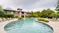 Located at 1448 Grove Park Dr in Columbus, Georgia, this impressive outdoor recreational space centers around a generously sized freeform swimming pool with a smooth, curved design that complements the natural surroundings. The pool is bordered by an expansive stamped concrete deck finished in warm earth tones, providing ample space for lounging and entertaining. Rows of sling-style chaise lounge chairs are arranged along the pool's perimeter, offering comfortable spots to relax and take in the views. The deck's organic, flowing shape follows the contours of the pool and surrounding greenery, creating a seamless transition between hardscape and landscape. Lush, mature landscaping frames the entire area, featuring towering magnolia trees, crape myrtles, tall pines, and carefully maintained mulched garden beds filled with ornamental shrubs and ground cover. A wrought iron perimeter fence with stone pillar accents provides security while maintaining an open, park-like aesthetic. A covered structure is visible at one end of the space, suggesting a pavilion or cabana for additional shade and amenities. The combination of natural greenery, thoughtfully designed hardscaping, and resort-style furnishings gives this outdoor space a serene, upscale ambiance.