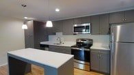 This single-floor space showcases a thoughtfully designed kitchen that serves as a central highlight. The kitchen features a cohesive aesthetic defined by flat-front gray cabinetry that extends floor-to-ceiling, providing generous storage throughout. Crisp white countertops offer a clean contrast against the darker cabinetry, while a matching white subway tile backsplash ties the look together seamlessly.

A full suite of stainless steel appliances includes an electric range, over-the-range microwave, and dishwasher, lending a polished, contemporary feel. The undermount sink is positioned along the perimeter countertop beneath recessed cabinetry, complemented by a modern brushed-nickel faucet.

A kitchen island with a white countertop surface provides additional prep space and casual seating, anchoring the layout and enhancing the kitchen's functionality. A single pendant light hangs above, adding focused task lighting while contributing to the overall modern ambiance.

Light hardwood flooring runs throughout, brightening the space and adding warmth that balances the cool tones of the gray cabinetry. Recessed ceiling lighting further enhances the clean, well-lit atmosphere of this updated kitchen.