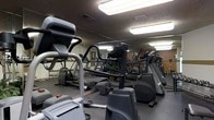 This fitness center is designed to support a comprehensive workout experience within a single-floor layout. The space is outfitted with a wide array of exercise equipment, including treadmills, elliptical machines, stair climbers, and a dedicated free weights area with a full rack of dumbbells. Floor-to-ceiling mirrors line the walls, visually expanding the room while allowing users to monitor their form during workouts. The ceiling features a textured finish and is illuminated by large, bright fluorescent light panels that ensure the space is well-lit throughout. The dark, neutral color palette of the equipment and flooring gives the gym a sleek, professional atmosphere. The thoughtful arrangement of machines allows for efficient use of the available floor space, providing ample room to move between stations. Overall, this fitness center offers a focused and functional environment suited for a range of fitness routines, from cardiovascular training to strength and resistance work.