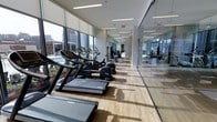 This fitness center is a well-appointed, single-floor facility designed to provide a comprehensive workout environment. The space is lined with floor-to-ceiling windows along one side, flooding the interior with natural light and offering sweeping views of the surrounding cityscape. The hardwood-style flooring adds a warm, refined aesthetic that contrasts with the sleek, modern equipment throughout.

The cardio zone is prominently featured, housing a row of treadmills, elliptical machines, and stationary bikes — all equipped with individual digital displays. The equipment is arranged to take full advantage of the exterior views, enhancing the workout experience. Recessed linear lighting runs along the ceiling, supplementing the natural light and maintaining a bright, energizing atmosphere throughout the day and evening.

A full-length glass wall partitions a separate workout area, visible from the main floor and offering additional training space with strength equipment. The transparent divider maintains an open, expansive feel while defining distinct zones within the facility. The overall design is clean and contemporary, prioritizing both functionality and visual appeal.