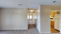 This single-story space is defined by a long, carpeted central hallway that serves as the primary circulation corridor connecting the various rooms. The hallway is lined with neutral-toned beige walls and features white baseboards and trim, creating a clean, consistent aesthetic throughout. Overhead, globe-style ceiling light fixtures are spaced along the corridor, providing warm, ambient illumination that complements the natural light filtering in through a window at the far end of the hallway. The textured ceiling adds a subtle visual element while housing practical HVAC vents for climate control. White panel doors with standard hardware are positioned along the hallway, offering access to individual rooms. The wall-to-wall carpet flooring in a muted brown tone runs the length of the corridor, adding warmth underfoot. The depth of the hallway gives the space a sense of length and connectivity, while the neutral color palette throughout maintains a cohesive and understated look. Light switch plates are evenly distributed along the walls, ensuring practical accessibility. Overall, the space presents a straightforward, functional layout with a calm and neutral ambiance.