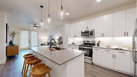 This single-floor residence offers a thoughtfully designed layout that maximizes functionality within a compact footprint. The open-concept kitchen and living area serves as the heart of the home, where a large quartz-topped island with an undermount sink and seating for multiple bar stools anchors the space. The kitchen features floor-to-ceiling white shaker cabinetry, a classic subway tile backsplash, and a full suite of stainless steel appliances, including a range, over-the-range microwave, and French door refrigerator. Pendant lighting with clear glass shades adds a modern touch overhead. The adjoining living area is finished with light wood-style flooring, a ceiling fan, and French doors that invite natural light. The bedroom provides a comfortable retreat, carpeted for warmth, with a ceiling fan, a window dressed with blinds and curtains, and access to a dedicated walk-in closet featuring dual hanging rods and built-in shelving. A hallway connects the private and shared spaces, contributing to an efficient, well-organized flow throughout the residence. The overall aesthetic is clean and contemporary, defined by a neutral palette and modern fixtures.