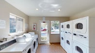 The laundry facility at 1845 N Broadway in Escondido is a practical, well-equipped space designed to meet residents' everyday needs. The single-floor room features tile flooring throughout, providing a clean and durable surface that is easy to maintain. Recessed ceiling lights offer consistent overhead illumination, supplemented by natural light streaming in through two windows and a glass-panel exterior door, which also provides a view of the surrounding outdoor area and contributes to an open, airy feel.

Along the left wall, a row of front-loading washing machines is neatly arranged side by side, each equipped with individual controls for user convenience. On the right side of the room, stacked dryer units maximize the use of vertical space, offering multiple drying options within a compact footprint. The card or coin-operated payment panels on the machines indicate a shared-use setup suited for a residential community.

A fire extinguisher is visibly mounted on the wall, reflecting attention to safety standards. Informational signage is posted near the entrance, keeping users informed of facility guidelines. The neutral beige walls and clean layout give the space a functional yet tidy ambiance.