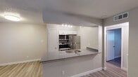 This single-floor unit is designed with a practical, open layout that connects the kitchen and living areas seamlessly. The kitchen is a standout feature, equipped with stainless steel appliances including a refrigerator, range with microwave hood, and a peninsula countertop finished in gray laminate. White cabinetry provides ample storage, while track lighting illuminates the workspace. A single-basin sink is integrated into the peninsula, which also functions as a casual dining or workspace surface.

The living area is generously sized, featuring light-toned wood-look flooring and a textured ceiling. Windows fitted with horizontal blinds allow natural light to filter in, creating a bright and open atmosphere. A ceiling fan serves the bedroom, which also features the same wood-look flooring in a slightly deeper tone, a window with blinds, and access to a closet.

The bathroom includes a combined tub and shower unit with tile surround, a toilet, and a vanity with a gray countertop and a framed mirror. Textured walls are consistent throughout the unit, complementing the neutral, warm color palette. The overall design is clean and functional, with consistent finishes tying the space together cohesively.