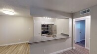 This single-floor unit is designed with a practical, open layout that connects the kitchen and living areas seamlessly. The kitchen is a standout feature, equipped with stainless steel appliances including a refrigerator, range with microwave hood, and a peninsula countertop finished in gray laminate. White cabinetry provides ample storage, while track lighting illuminates the workspace. A single-basin sink is integrated into the peninsula, which also functions as a casual dining or workspace surface.

The living area is generously sized, featuring light-toned wood-look flooring and a textured ceiling. Windows fitted with horizontal blinds allow natural light to filter in, creating a bright and open atmosphere. A ceiling fan serves the bedroom, which also features the same wood-look flooring in a slightly deeper tone, a window with blinds, and access to a closet.

The bathroom includes a combined tub and shower unit with tile surround, a toilet, and a vanity with a gray countertop and a framed mirror. Textured walls are consistent throughout the unit, complementing the neutral, warm color palette. The overall design is clean and functional, with consistent finishes tying the space together cohesively.