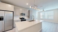 This single-floor residence is designed with a clean, contemporary aesthetic that emphasizes bright, open living. The kitchen serves as the centerpiece of the home, showcasing an expansive quartz island with an undermount sink and a brushed nickel pull-down faucet. A sleek pendant light fixture illuminates the island from above. Soft gray shaker-style cabinetry lines the walls alongside white subway tile backsplash, while a full suite of stainless steel appliances—including a side-by-side refrigerator with ice maker, electric range, and built-in microwave—provides modern functionality. Light wood-look plank flooring extends through the kitchen and main living areas, adding warmth to the neutral palette.

The living area benefits from large windows fitted with horizontal blinds, allowing natural light to fill the space. Bedrooms feature plush carpet flooring, ceiling fans, and generous windows overlooking greenery outside. The bathroom is finished with gray cabinetry, a white quartz vanity top, light wood-look tile flooring, and a bathtub with grab bar. A dedicated laundry room houses a full-sized washer and dryer with overhead wire shelving, rounding out the practical, well-appointed layout.