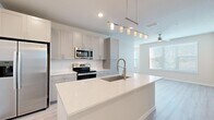 This single-floor residence is designed with a clean, contemporary aesthetic that emphasizes bright, open living. The kitchen serves as the centerpiece of the home, showcasing an expansive quartz island with an undermount sink and a brushed nickel pull-down faucet. A sleek pendant light fixture illuminates the island from above. Soft gray shaker-style cabinetry lines the walls alongside white subway tile backsplash, while a full suite of stainless steel appliances—including a side-by-side refrigerator with ice maker, electric range, and built-in microwave—provides modern functionality. Light wood-look plank flooring extends through the kitchen and main living areas, adding warmth to the neutral palette.

The living area benefits from large windows fitted with horizontal blinds, allowing natural light to fill the space. Bedrooms feature plush carpet flooring, ceiling fans, and generous windows overlooking greenery outside. The bathroom is finished with gray cabinetry, a white quartz vanity top, light wood-look tile flooring, and a bathtub with grab bar. A dedicated laundry room houses a full-sized washer and dryer with overhead wire shelving, rounding out the practical, well-appointed layout.