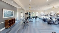 Located at 4255 Kittredge St in Denver, CO, this single-floor fitness center is thoughtfully designed to support a wide range of workout routines. The facility is organized into distinct zones, each serving a specific fitness purpose while maintaining a cohesive, modern aesthetic throughout.

The main cardio area is a standout feature, lined with treadmills, elliptical machines, and a stationary bike positioned in front of a striking row of arched windows. These windows frame views of an outdoor pool and surrounding greenery, filling the space with natural light and creating an energizing atmosphere. Ceiling fans and recessed lighting complement the bright, airy feel.

Adjacent to the cardio zone is a strength training area equipped with a cable machine, a leg press, and a dedicated free weights station featuring a full rack of kettlebells and dumbbells. A wall-mounted clock and flat-screen TV add functional touches to the space, which features durable wood-look flooring and crown molding for a polished finish.

A separate studio room provides an open, versatile space for functional training, featuring a large wall mirror, stability balls, a freestanding punching bag, and the same arched windows that offer views of the landscaped grounds outside. The overall design balances practicality with an inviting ambiance, making this fitness center a comprehensive and well-appointed facility.