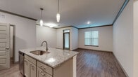 Located at 114 SE Douglas St in Lee's Summit, Missouri, this well-appointed single-floor residence combines modern finishes with a practical, open layout. The kitchen serves as a central highlight, anchored by a large granite-topped island with an undermount sink and pendant lighting overhead. White shaker-style cabinetry, a subway tile backsplash, and a full suite of stainless steel appliances — including a refrigerator, range, and over-the-range microwave — complete the space. An in-unit stacked washer and dryer is conveniently accessible just off the kitchen. The open-concept living and dining area features rich hardwood-style plank flooring, crown molding, and generous natural light streaming through multiple windows with blinds, creating a bright and welcoming atmosphere. A glass-paneled entry door adds additional light to the main living space. The bedrooms offer plush carpeting, ample wall space, and windows that bring in natural light. The bathroom is finished with a dual-sink granite vanity, a large wall mirror with overhead lighting, and hardwood-style flooring, reflecting the same quality of finishes found throughout the home. The neutral color palette and contemporary detailing give the entire space a clean, move-in-ready appeal.