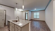 Located at 114 SE Douglas St in Lee's Summit, Missouri, this well-appointed single-floor residence combines modern finishes with a practical, open layout. The kitchen serves as a central highlight, anchored by a large granite-topped island with an undermount sink and pendant lighting overhead. White shaker-style cabinetry, a subway tile backsplash, and a full suite of stainless steel appliances — including a refrigerator, range, and over-the-range microwave — complete the space. An in-unit stacked washer and dryer is conveniently accessible just off the kitchen. The open-concept living and dining area features rich hardwood-style plank flooring, crown molding, and generous natural light streaming through multiple windows with blinds, creating a bright and welcoming atmosphere. A glass-paneled entry door adds additional light to the main living space. The bedrooms offer plush carpeting, ample wall space, and windows that bring in natural light. The bathroom is finished with a dual-sink granite vanity, a large wall mirror with overhead lighting, and hardwood-style flooring, reflecting the same quality of finishes found throughout the home. The neutral color palette and contemporary detailing give the entire space a clean, move-in-ready appeal.