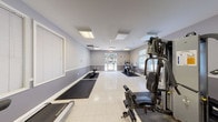 This fitness center offers a spacious, single-floor layout designed to accommodate a variety of workout routines. The interior is finished with two-tone gray walls accented by white chair rail molding, and speckled tile flooring throughout, lending the space a clean and functional aesthetic. Flush-mounted ceiling light panels provide consistent, even illumination across the room.

Natural light enters through multiple windows fitted with horizontal blinds, offering views of surrounding greenery. A glass-paneled exterior door at one end of the room further enhances the bright atmosphere while providing convenient direct access to the outdoors.

The fitness area is well-appointed with a range of equipment, including a treadmill, an elliptical trainer, a stationary bike, and a multi-station cable weight machine. Rubber exercise mats are positioned on the floor to define workout zones and protect the flooring. A weight bench and additional free-weight equipment round out the strength training options.

A short entry corridor leads into the main fitness room, where a wall-mounted water fountain provides convenient hydration access. Signage identifies the space as a Fitness Center, and a fire alarm panel is visible near the exterior door, reflecting standard safety compliance. The overall ambiance is practical and welcoming, suited for daily exercise use.