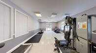 This fitness center offers a spacious, single-floor layout designed to accommodate a variety of workout routines. The interior is finished with two-tone gray walls accented by white chair rail molding, and speckled tile flooring throughout, lending the space a clean and functional aesthetic. Flush-mounted ceiling light panels provide consistent, even illumination across the room.

Natural light enters through multiple windows fitted with horizontal blinds, offering views of surrounding greenery. A glass-paneled exterior door at one end of the room further enhances the bright atmosphere while providing convenient direct access to the outdoors.

The fitness area is well-appointed with a range of equipment, including a treadmill, an elliptical trainer, a stationary bike, and a multi-station cable weight machine. Rubber exercise mats are positioned on the floor to define workout zones and protect the flooring. A weight bench and additional free-weight equipment round out the strength training options.

A short entry corridor leads into the main fitness room, where a wall-mounted water fountain provides convenient hydration access. Signage identifies the space as a Fitness Center, and a fire alarm panel is visible near the exterior door, reflecting standard safety compliance. The overall ambiance is practical and welcoming, suited for daily exercise use.
