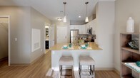 Located at 11 Bancroft in Nashua, New Hampshire, this single-floor residence offers a thoughtfully designed layout that balances comfort and modern style. The open-concept kitchen is a standout feature, showcasing white cabinetry, stainless steel appliances including a refrigerator, microwave, and range, as well as a warm-toned countertop island with an undermount sink and bar seating — ideal for casual dining and entertaining. Light wood-style flooring flows throughout, complementing the neutral, taupe-toned walls that lend the space a calm, cohesive ambiance. The living room is generously proportioned, filled with natural light from large windows dressed in patterned drapes and roller shades, creating a welcoming and airy atmosphere. The space accommodates a comfortable seating arrangement and media center with ease. Two well-sized bedrooms each feature closets, natural light from windows, and a tranquil neutral palette. The primary bathroom is finished with dark cabinetry, a white quartz-style countertop, open shelving, and direct access to an in-unit laundry area. A second full bathroom includes a combined tub and shower, a vanity with dark cabinetry, and tile flooring. The overall design is clean, contemporary, and move-in ready.