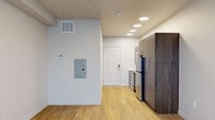 This single-floor unit offers a clean, modern design highlighted by light hardwood-style flooring that runs consistently throughout the space, creating a cohesive and warm aesthetic. The kitchen is efficiently laid out and features stainless steel appliances, including a refrigerator, electric range, and over-the-range microwave, all complemented by rich wood-toned cabinetry that extends to the ceiling for ample storage. Recessed lighting lines the kitchen ceiling, providing bright, even illumination. The entry area is practical and well-organized, housing a thermostat and an electrical panel flush against the wall. Adjacent to the kitchen, the open living area is bathed in natural light from a window fitted with horizontal blinds, offering a versatile space with a neutral color palette and recessed lighting overhead. The bathroom includes a full-size soaking tub with a shower combination, built-in corner shelves, and classic white finishes throughout. Clean white walls, efficient HVAC ventilation, and a keypad-secured front entry door round out the thoughtfully designed interior, making the most of every square foot in a functional and contemporary way.