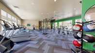 This single-floor fitness center offers a comprehensive workout facility designed to support a wide range of exercise routines. The main workout area is expansive and airy, featuring high ceilings and large floor-to-ceiling windows along multiple walls that flood the space with natural light and provide views of the surrounding landscaped grounds. A bold green accent wall adds a vibrant, energetic atmosphere throughout.

The flooring throughout the facility is a stylish herringbone-patterned carpet tile in neutral tones, lending both comfort and visual appeal underfoot. The main floor is comprehensively equipped with treadmills, elliptical machines, stair climbers, a rowing machine, and stationary bikes for cardio training. A dedicated free weights section features a full rack of dumbbells, weight benches, and cable machines, while a central power rack structure supports functional training with TRX suspension trainers and battle ropes. A wall of mirrors enhances the sense of space in the strength training zone.

A separate studio room, distinguished by its own green accent wall and rustic wood shelving unit, provides a flexible space for group fitness, stretching, or yoga, with low workout benches and wall-mounted mirrors. Near the entry, a water fountain and sanitizing station offer convenience. Recessed lighting and ceiling fans ensure a comfortable environment throughout the facility.