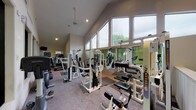 This single-floor fitness center is thoughtfully designed to support a comprehensive workout experience. The space is defined by its striking vaulted ceilings, which rise dramatically overhead and are lined with a continuous row of large windows along one side, flooding the interior with natural light and offering views of the surrounding greenery outside. Recessed lighting and pendant fixtures provide additional illumination throughout.

The main workout area is generously sized and features a wide selection of strength training equipment, including multiple Paramount Fitnessline cable and weight machines, adjustable benches, and a dedicated free-weight station. A row of floor-to-ceiling mirrors runs along one wall, enhancing the sense of space and allowing users to monitor their form during exercise.

Cardio equipment is well-represented, with treadmills, an elliptical trainer, and an upright stationary bike positioned throughout the room. Rubber matting beneath select machines protects the neutral-toned carpet flooring. A wall-mounted flat-screen television is positioned near the cardio area for entertainment during workouts.

A separate windowed room, visible through interior glass panels, provides an additional dedicated workout zone with its own equipment and bright lighting. The overall ambiance is clean, open, and functional, making this fitness center a well-rounded facility suitable for a full range of exercise routines.