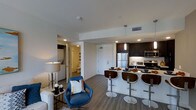 Located at 100 Citronica in Lemon Grove, this single-floor residence offers a thoughtfully designed open-concept layout that seamlessly connects the living and kitchen areas. The kitchen is a standout feature, outfitted with dark espresso cabinetry, stainless steel appliances including a refrigerator, microwave, and range, and a sleek kitchen island with modern bar stools that provide casual seating. Pendant lighting above the island adds a contemporary touch, while recessed lighting throughout keeps the space bright and welcoming. The living area complements the kitchen with hardwood-style flooring that flows cohesively throughout the space, creating a warm and unified aesthetic. A vibrant blue accent chair adds a pop of color and personality to the sitting area. Crisp white walls and clean architectural lines contribute to a modern, airy ambiance. Down the hallway, an in-unit laundry area offers added convenience, alongside a full bathroom visible from the corridor. The layout is efficient yet spacious, making excellent use of every square foot while maintaining a comfortable and functional living environment throughout.
