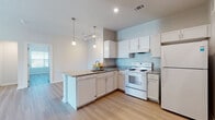 This single-story unit located along SH-123 in San Marcos, Texas offers a clean, modern interior with a cohesive design throughout. Light wood-tone flooring runs continuously across the main living areas and bedrooms, creating a bright and unified aesthetic that complements the neutral white and soft gray wall palette.

The kitchen serves as a central feature, equipped with white shaker-style cabinetry, granite countertops, and a peninsula with an integrated sink and dishwasher. A full suite of white appliances includes an electric range and refrigerator. Pendant lighting hangs above the peninsula, while recessed lighting provides additional illumination. The kitchen opens directly to the living and dining area, which benefits from natural light and sliding glass doors leading to a private balcony with exterior views.

The bedrooms are generously proportioned, each featuring ceiling fans and ample closet space, including at least one room with a built-in closet shelving system. Bathrooms are finished with stone-look tile flooring, white vanity cabinetry, and a full tub and shower combination. At least one bedroom includes a connected bathroom. The overall layout is efficient and well-lit, with a calm, residential ambiance throughout.