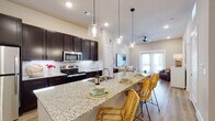 This single-floor residence offers a thoughtfully designed open-concept layout where the kitchen, dining, and living areas flow seamlessly together. The kitchen is a standout feature, showcasing rich dark cabinetry that reaches toward the ceiling, a herringbone-patterned tile backsplash, and a full suite of stainless steel appliances. A generously sized granite-topped island serves as the centerpiece, offering bar seating for multiple guests beneath a trio of elegant glass pendant lights — ideal for both casual dining and entertaining.

The living area extends naturally from the kitchen, bathed in natural light through a set of glass-paned French doors. A ceiling fan adds comfort, while light-toned hardwood-style flooring runs consistently throughout, unifying the space with warmth and continuity.

The bedroom is well-proportioned, featuring a large window that allows ample natural light, and connects to a bathroom equipped with dark cabinetry, granite countertops, a large mirror, and hardwood-style flooring. The bathroom also features a shower with a classic white subway tile surround.

Throughout the residence, neutral warm-toned walls and recessed lighting create a bright, welcoming ambiance that complements the modern yet approachable design aesthetic.