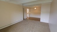 This single-floor space features an open-concept layout that connects the living area, dining space, and kitchen in a cohesive flow. The living room is finished with neutral-toned carpet and is framed by exposed ceiling beams that add a subtle architectural detail to the otherwise clean, minimalist aesthetic. A baseboard heating unit runs along the perimeter wall, providing an efficient heating solution.

The space transitions into a dining area distinguished by hardwood flooring, creating a clear visual separation between the two zones without the need for walls. A pendant light hangs above this area, offering focused ambient lighting that anchors the dining space. The neutral wall tones throughout contribute to a bright and open atmosphere.

Adjacent to the dining area, the kitchen is accessible through an open pass-through counter with white cabinetry visible from the main living space. The kitchen layout appears functional and compact, with appliances integrated neatly into the design. The consistent use of neutral tones across all areas creates a cohesive and understated interior that is versatile and move-in ready.