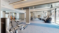 Located at 7800 E Peakview Ave in Centennial, Colorado, this fitness center offers an impressively spacious exercise environment spread across a single floor. The facility's open layout is anchored by structural columns that define distinct workout zones without interrupting the overall sense of flow and openness. Large floor-to-ceiling windows line one side of the space, flooding the interior with abundant natural light and providing views of the surrounding area, while ceiling fans and recessed lighting ensure a comfortable environment throughout. The patterned carpet flooring — featuring a subtle geometric design in cool blue and grey tones — adds a polished, modern aesthetic underfoot. Full-length mirrors are strategically positioned to amplify the sense of space and support training. Equipment is thoughtfully distributed across zones: cardio machines including treadmills, stair climbers, rowing machines, and stationary bikes occupy one section, while strength training areas feature cable machines, weight benches, and a well-stocked free weights section with kettlebells, dumbbells, and stability balls. A dedicated open stretch and functional training area provides room for dynamic movement. A wall-mounted television, ceiling fans, and private restroom access round out the amenities, making this a fully equipped, professional-grade fitness facility.