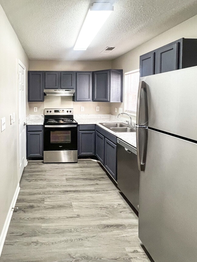 Vista de la cocina tipo galera con refrigerador de acero inoxidable, lavavajillas, gabinetes oscuros, encimeras estilo mármol y pisos con apariencia de madera. - Pendleton Townhomes
