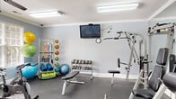 Located at 3401 Bremer Hall Ct in Raleigh, NC, this fitness center is a fully equipped, single-room facility designed to support a wide range of workout routines. The space features soft blue-gray walls, white crown molding, and a durable rubber-floored surface ideal for high-traffic athletic use. Dual ceiling fans and flush fluorescent lighting ensure the room remains well-ventilated and brightly lit throughout. Large windows fitted with white blinds allow natural light to filter in while maintaining privacy. A wall-mounted flat-screen television provides entertainment during workouts, accompanied by a wall clock for time management. The cardio zone includes treadmills, an elliptical trainer, and a stationary spin bike — all TRUE-brand machines. Strength training is supported by a multi-station cable machine, a weight bench, and a tiered dumbbell rack with a comprehensive range of free weights. Functional training accessories are well-represented, with a dedicated rack holding medicine balls, stability balls, resistance bands, and foam rollers. Full-length wall mirrors on one side of the room visually expand the space and assist with exercise form. A fire extinguisher and posted safety guidelines reflect a commitment to user safety. The entrance features double doors, providing convenient access to this comprehensive fitness facility.
