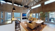 This clubhouse delivers a warm and sophisticated atmosphere through its striking architectural design. The centerpiece of the space is a dramatic vaulted ceiling supported by exposed timber trusses, which draws the eye upward and creates an expansive, airy feel throughout. Warm wood-paneled walls run the length of the interior, lending a natural, lodge-inspired character that is both refined and welcoming.

The open-concept layout seamlessly integrates a lounge and social area, anchored by a sleek linear fireplace set beneath a large wall-mounted television — an ideal focal point for gatherings. Comfortable mid-century modern seating, including a mustard-toned sofa and leather accent chairs arranged over a richly textured area rug, creates an inviting and stylish conversation area. Pendant lighting, including a sculptural globe fixture, adds ambient warmth alongside the recessed track lighting above.

A bar counter with wood-toned bar stools provides a casual seating and service area, perfect for entertaining. Generous windows and clerestory glazing along the upper walls allow abundant natural light to pour in throughout the day, while offering views of the surrounding outdoor spaces. The overall ambiance is relaxed yet polished, making this clubhouse a versatile and welcoming retreat for residents and guests alike.