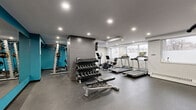 Located at 5501 Adalbert in Côte-Saint-Luc, QC, this fitness facility offers a thoughtfully designed workout space that caters to a wide range of exercise needs. The gym features a modern aesthetic defined by bold teal accent walls, textured hexagonal tile panels, and a neutral grey color palette throughout, creating an energizing yet functional atmosphere.

The space is outfitted with a strong selection of cardio equipment, including multiple treadmills and an elliptical machine, positioned alongside windows that allow natural light to fill the area. A robust multi-station cable machine, a barbell rack with weight plates, and a full tiered dumbbell rack provide comprehensive strength training options. Additional equipment includes a rowing machine, an adjustable weight bench, stability balls, and a wall-mounted flat-screen television.

Full-length mirrors line one wall, visually expanding the space and allowing users to monitor their form during workouts. Recessed lighting throughout ensures the gym is consistently and evenly illuminated. The durable rubber flooring provides safety and comfort underfoot. The facility is accessible via clearly marked entry doors and is equipped with emergency exit signage. Overall, this gym delivers a compact yet complete fitness environment designed to support diverse training routines.