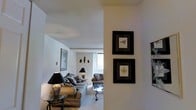 This single-floor residence offers a well-appointed and thoughtfully arranged living space with a clean, neutral aesthetic throughout. The living area is a standout feature, characterized by soft, light-toned walls and plush, cushioned seating that creates a warm and welcoming atmosphere. Natural light filters in through a generously sized window, brightening the room and highlighting the light-colored carpeting underfoot. Decorative table lamps with classic black shades add a refined touch and provide warm ambient lighting. The walls are adorned with framed botanical and nature-inspired artwork, lending a sophisticated yet approachable character to the space. A glass-topped coffee table anchors the seating arrangement, adding an element of elegance. Architectural detailing such as a structural column or partial wall creates a natural visual separation within the open layout, adding depth and dimension to the interior. The overall ambiance is calm and curated, with a cohesive design palette that balances comfort and style. This space is well-suited for relaxed everyday living while maintaining a polished and presentable appearance throughout.